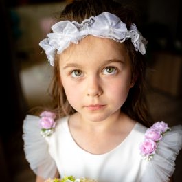 Dziewczynka z bukietem kwiatów ubrana w białą sukienkę i wianek na głowie.