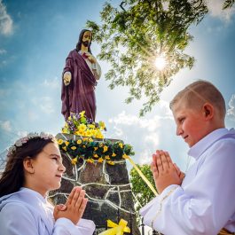 Pamiątka pierwszej komunii świętej - zdjęcie dzieci w albach pod przydrożną figurą Jezusa.